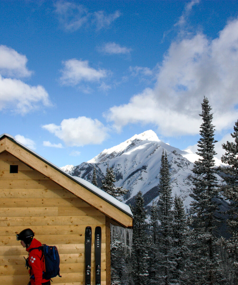 A skier with a red jacket stands by a backcountry cabin in Banffs snowy Rockies.