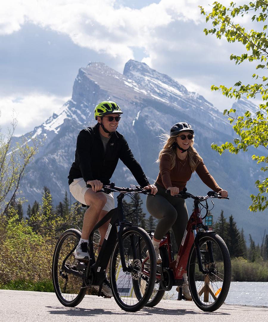 Two cyclists ride side by side on a scenic mountain road near Banff, Alberta.